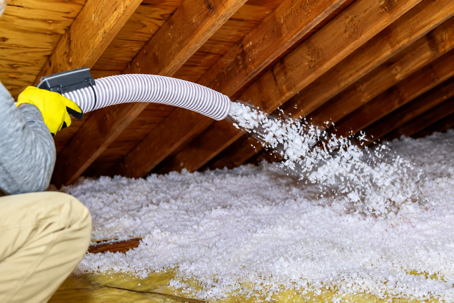 Worker directs a hose spraying loose white insulation across an attic floor beneath wooden roof rafters.