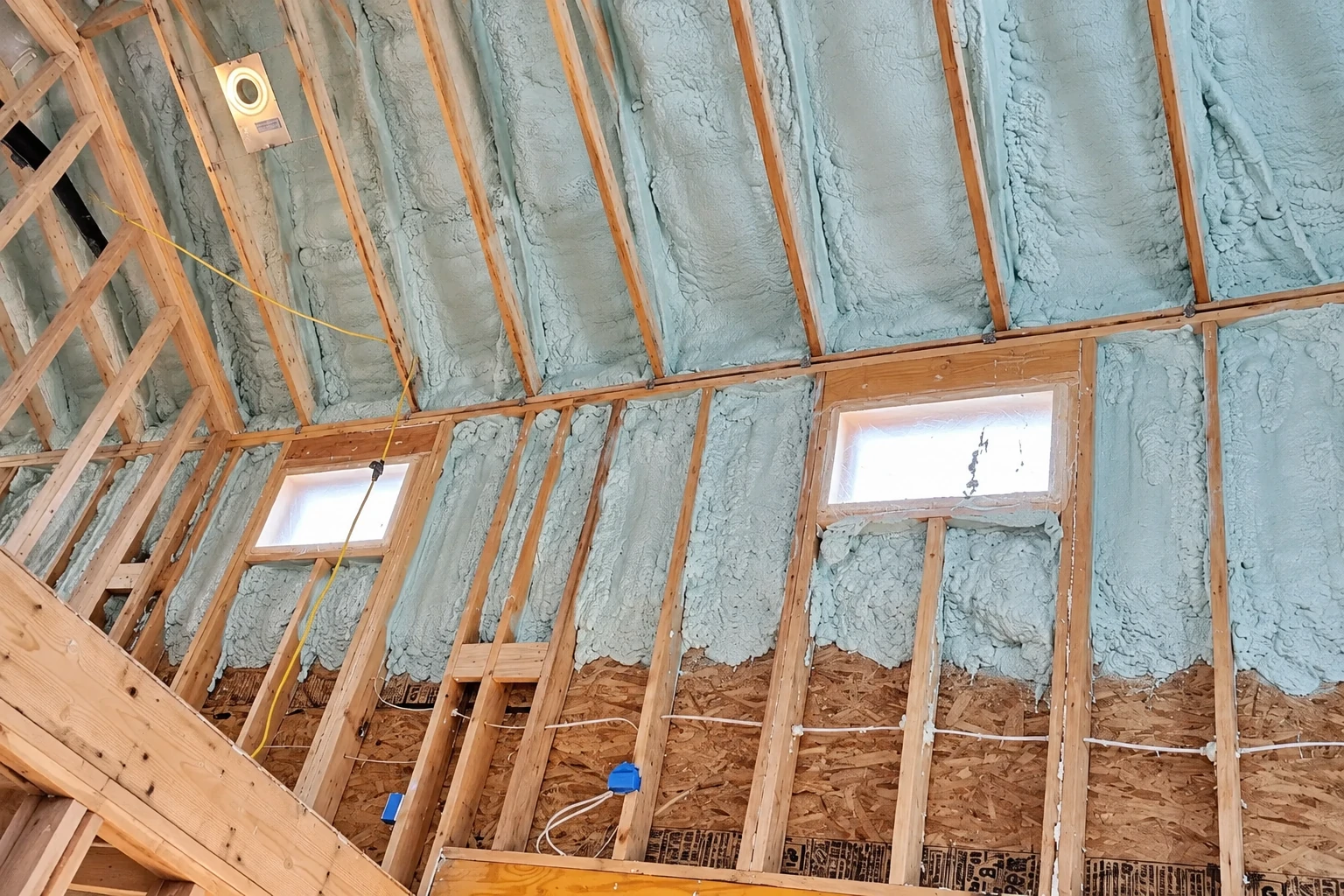 Blue-gray spray foam fills the roof framing around two small windows in an unfinished attic.