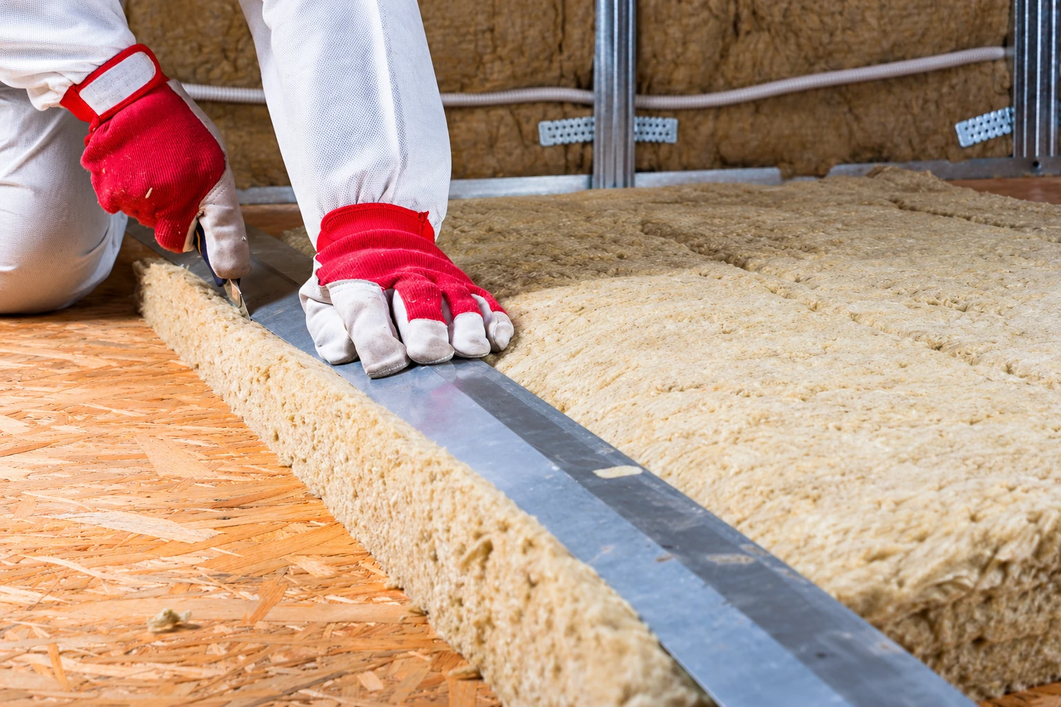 A worker in red gloves cuts a tan insulation batt to size with a straightedge on a wood subfloor.