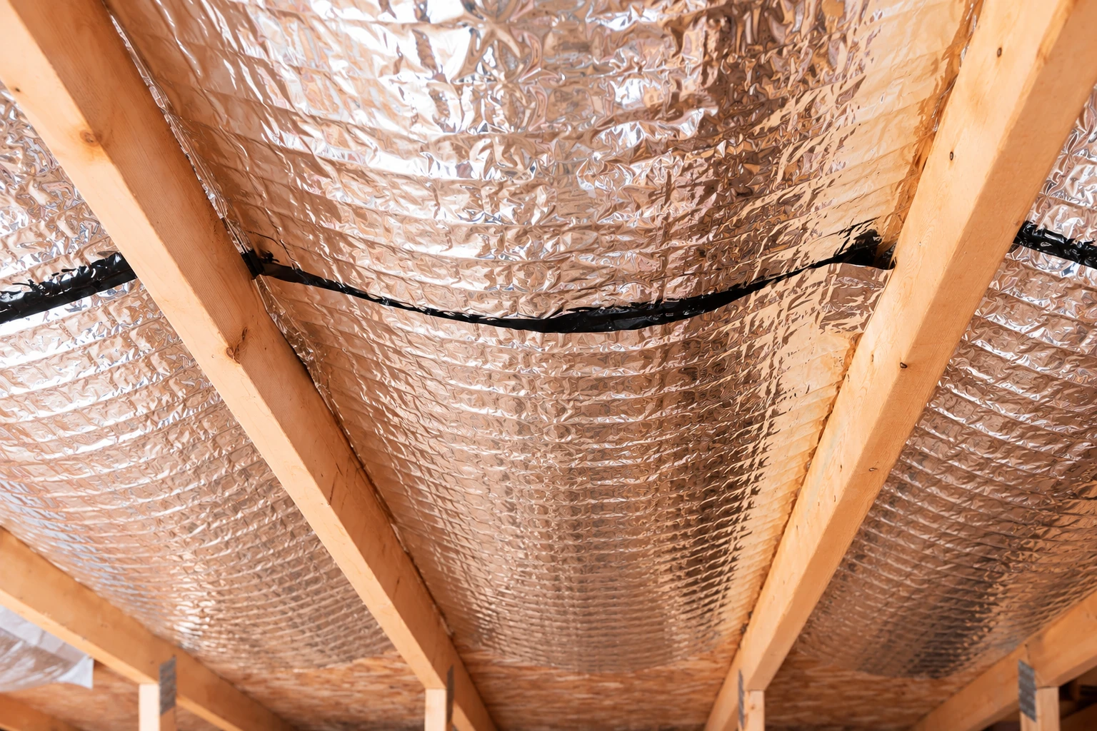 Reflective foil vapor barrier is stapled between wooden roof rafters.