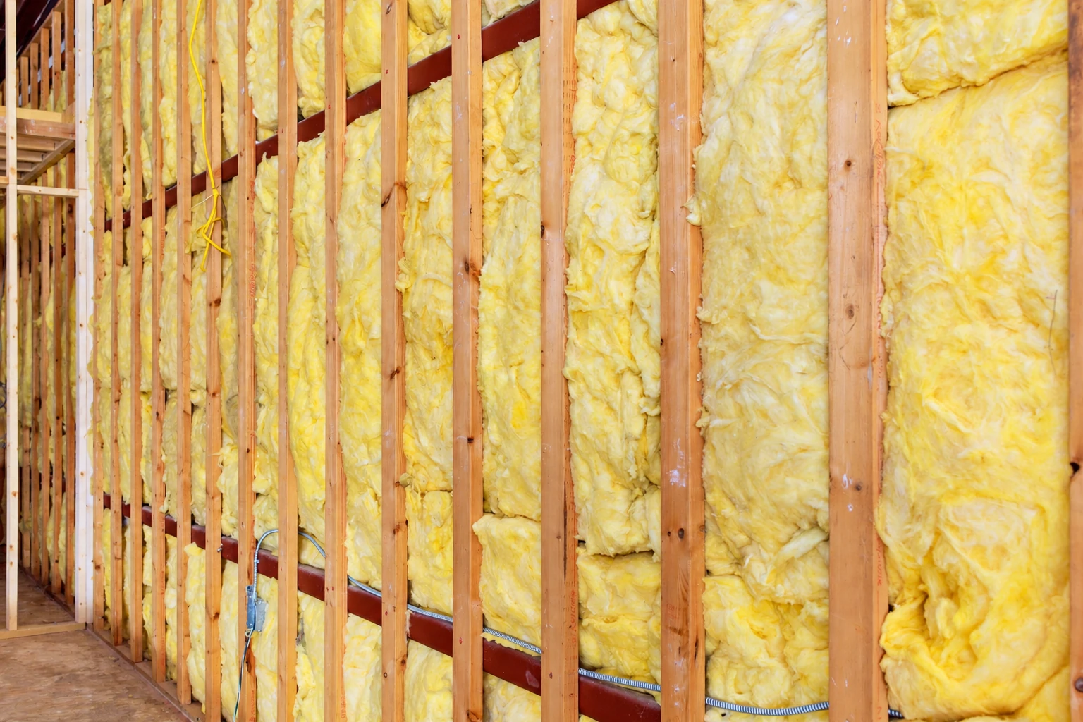 Yellow fiberglass batts fill the spaces between exposed wooden wall studs, with electrical wiring along the framing.